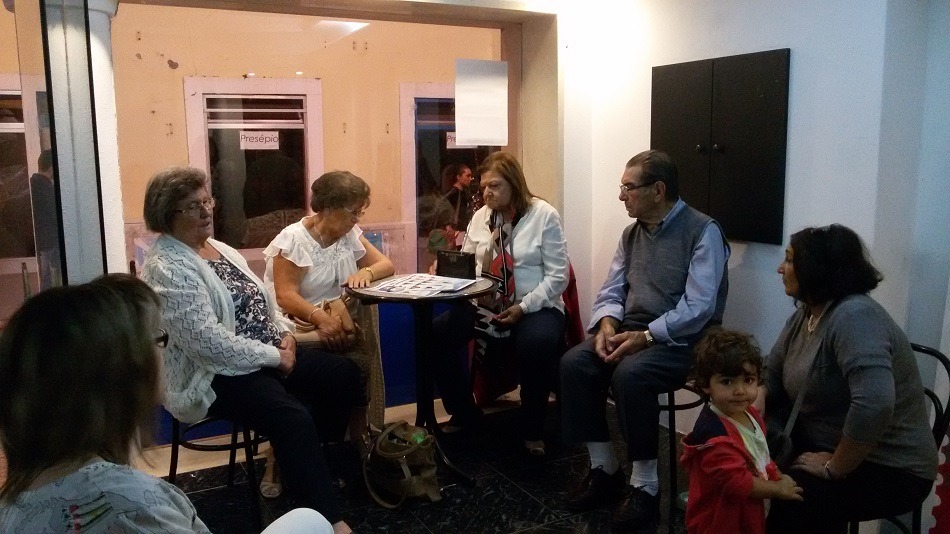 Um grupo de adultos e uma criança está reunido numa sala, junto a uma mesa com papéis. Ao fundo, vê-se uma porta com a indicação «Presépio».