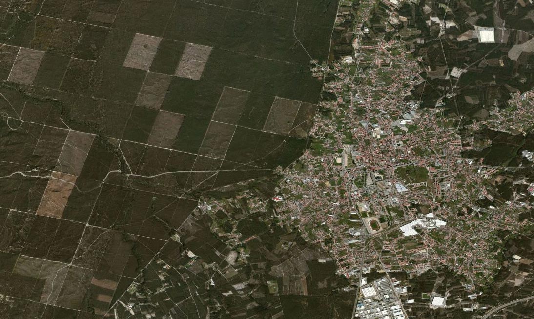 Vista aérea de uma localidade rodeada por áreas agrícolas e florestais. São visíveis ruas, edifícios e terrenos divididos em parcelas.