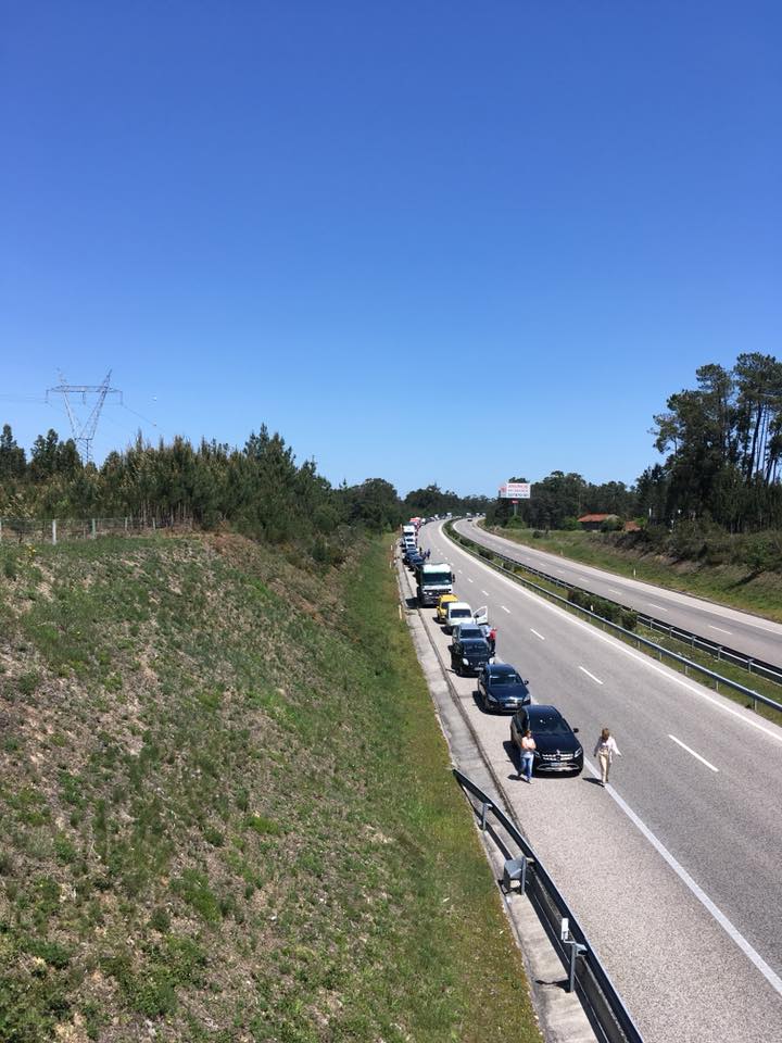 Uma fila de veículos está parada numa autoestrada. Duas pessoas caminham junto aos carros, numa faixa de rodagem.