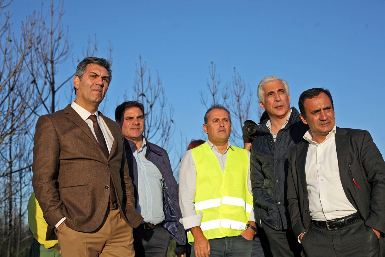 Um grupo de homens está reunido ao ar livre. Um deles veste um colete refletor amarelo; os restantes usam casacos ou fatos.