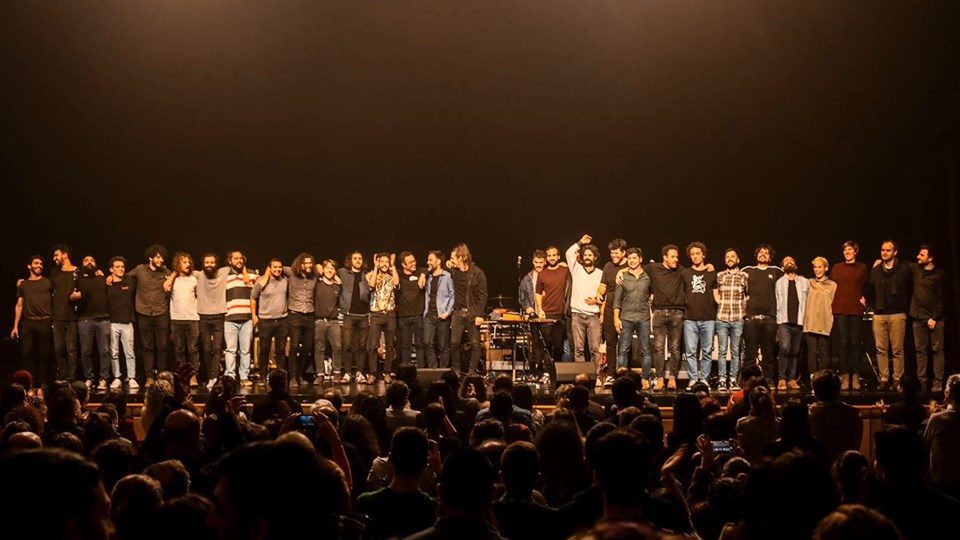 Um grupo numeroso de pessoas está alinhado num palco, diante de uma plateia. Algumas têm os braços sobre os ombros umas das outras e uma acena ao público.