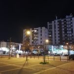 Vista noturna de uma praça e de uma avenida, com prédios de habitação, estabelecimentos iluminados e carros estacionados. Vêem-se algumas pessoas junto às lojas e às esplanadas.