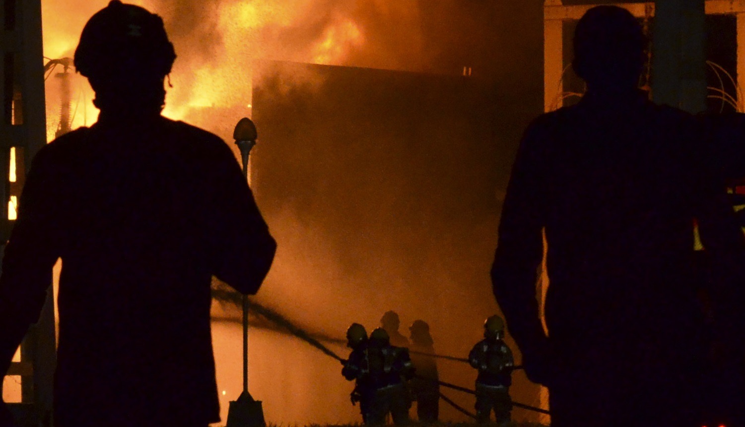 Várias pessoas, incluindo bombeiros com mangueiras, estão diante de um incêndio intenso. As chamas e o fumo iluminam a cena, enquanto duas figuras aparecem em primeiro plano, em silhueta.