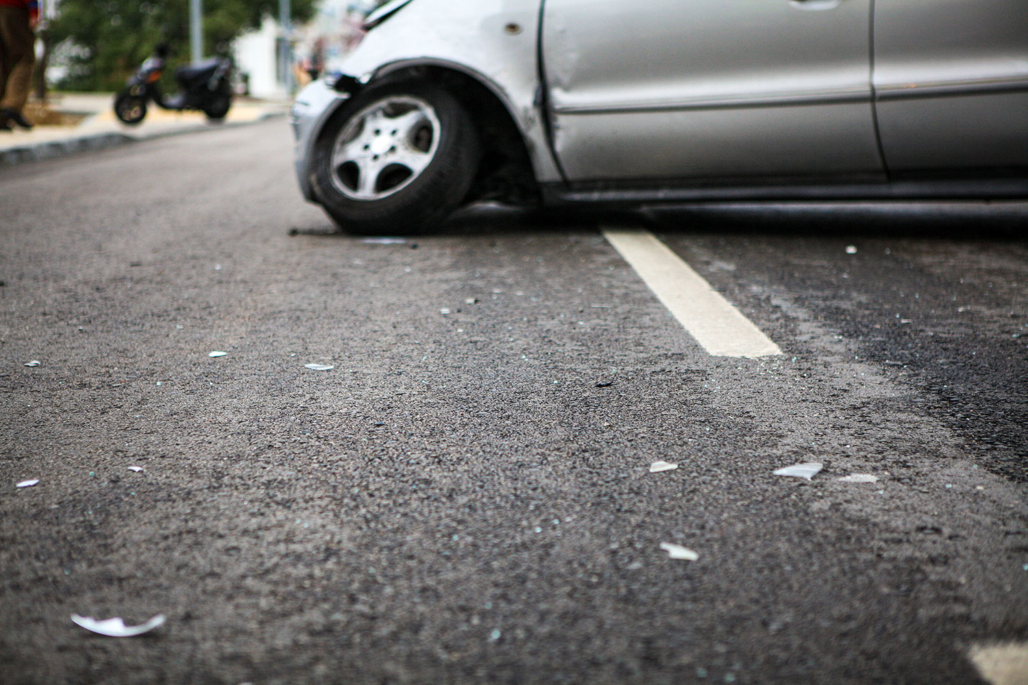 Um automóvel prateado está imobilizado na estrada com o pneu dianteiro vazio. Veem-se pequenos fragmentos espalhados pelo asfalto.