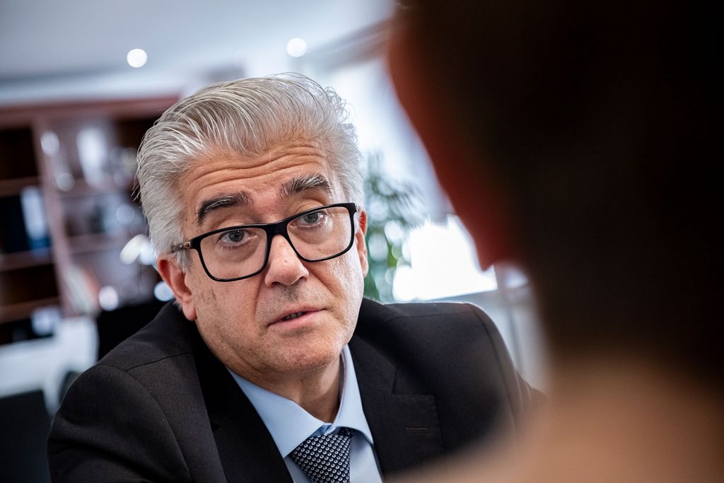 Um homem de cabelo grisalho, óculos e fato fala com outra pessoa num espaço interior. A imagem não contém texto visível que permita identificá-lo.