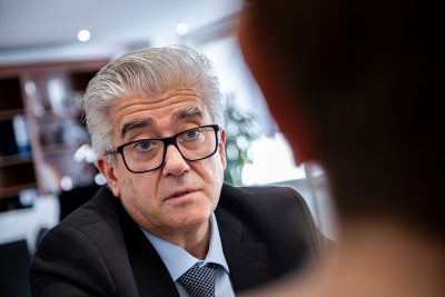 Um homem de cabelo grisalho, óculos e fato fala com outra pessoa num espaço interior. A imagem não contém texto visível que permita identificá-lo.