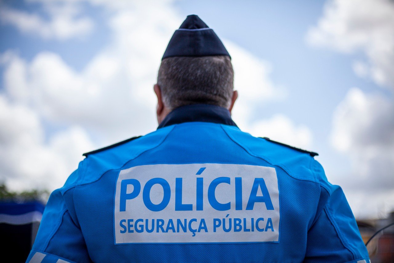 Um agente policial é fotografado de costas, com um uniforme azul onde se lê «Polícia Segurança Pública». O céu nublado ocupa o fundo da imagem.