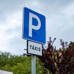 Um sinal azul de estacionamento, com a letra P, está sobre uma placa com a indicação «TÁXIS». Ao fundo veem-se árvores, um edifício e céu nublado.