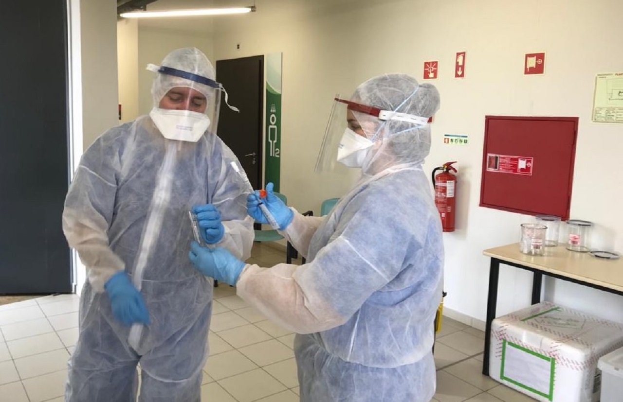 Duas pessoas com fatos de proteção, máscaras, viseiras e luvas manuseiam material numa sala. A imagem não permite determinar o procedimento em curso.