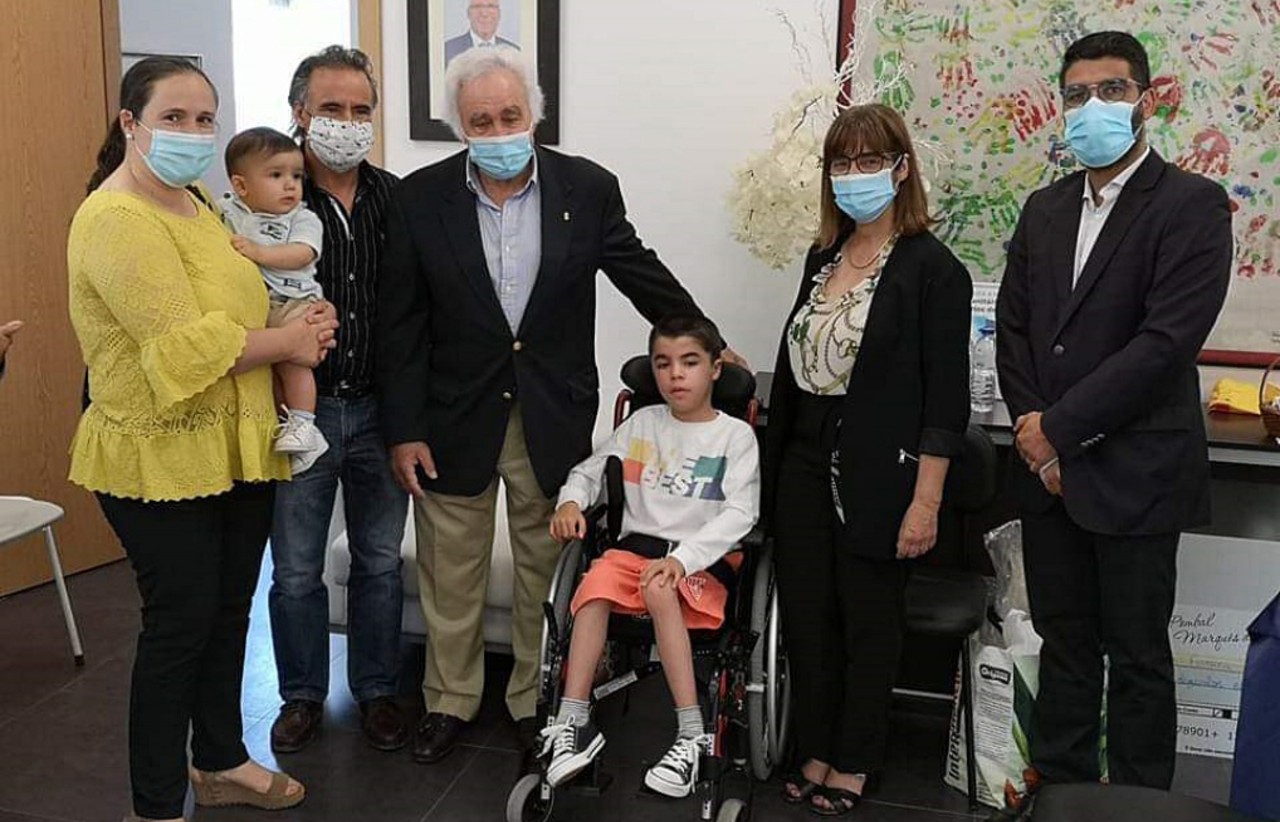 Seis pessoas e uma criança pequena posam numa sala; um jovem está numa cadeira de rodas. Vários adultos usam máscaras de proteção.
