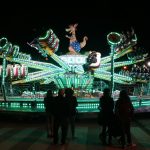 À noite, várias pessoas andam numa atração de feira iluminada com luzes verdes e brancas. Outras observam a atração em primeiro plano.
