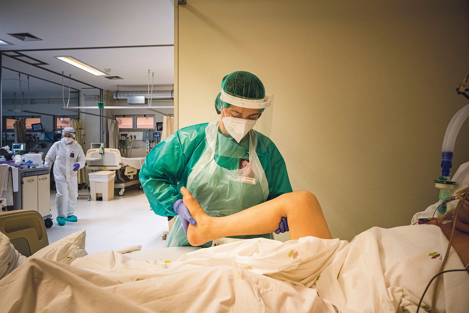 Fisioterapeuta com bata, máscara e viseira ajuda a movimentar a perna de um doente que está acamado