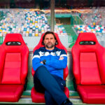 Um homem com roupa desportiva azul está sentado, de braços cruzados, num banco de suplentes com cadeiras vermelhas. Ao fundo vêem-se as bancadas de um estádio, com assentos de várias cores.