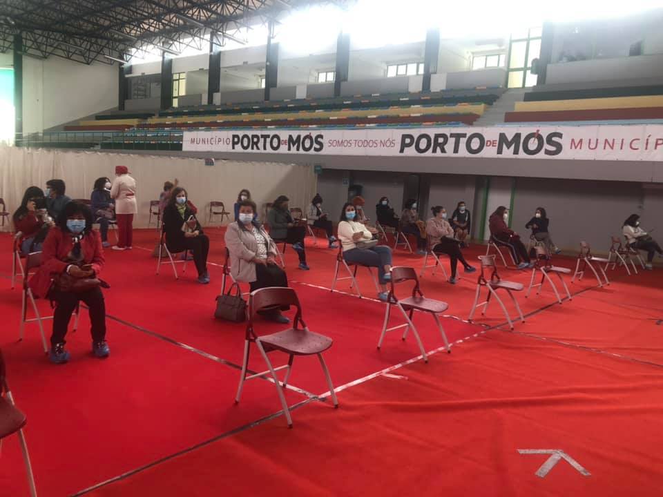 Pessoas com máscara estão sentadas em cadeiras espaçadas num pavilhão com piso vermelho. Nas bancadas, é visível a inscrição «Município de Porto de Mós».
