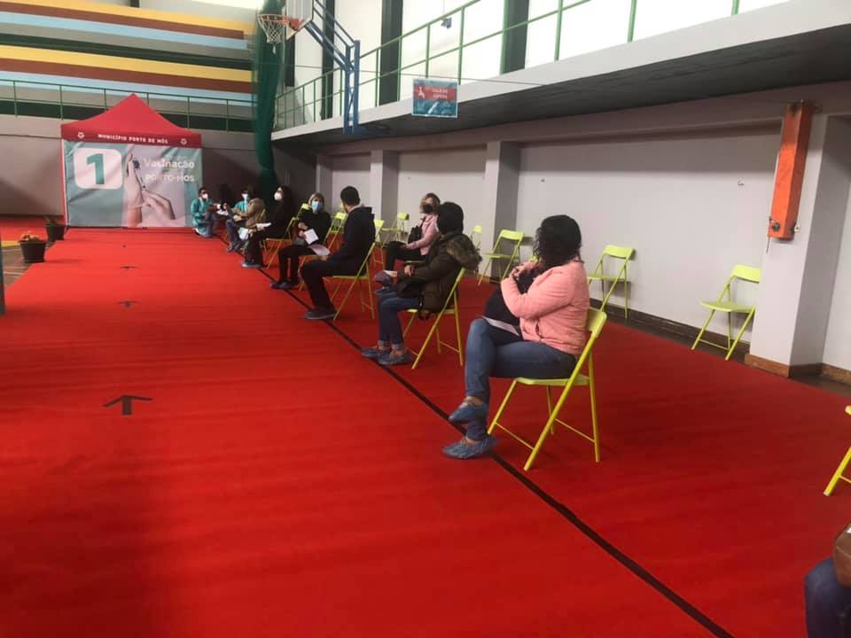 Num pavilhão desportivo, várias pessoas aguardam sentadas em cadeiras espaçadas, algumas com máscara. Ao fundo, há sinalética de vacinação contra a COVID-19.