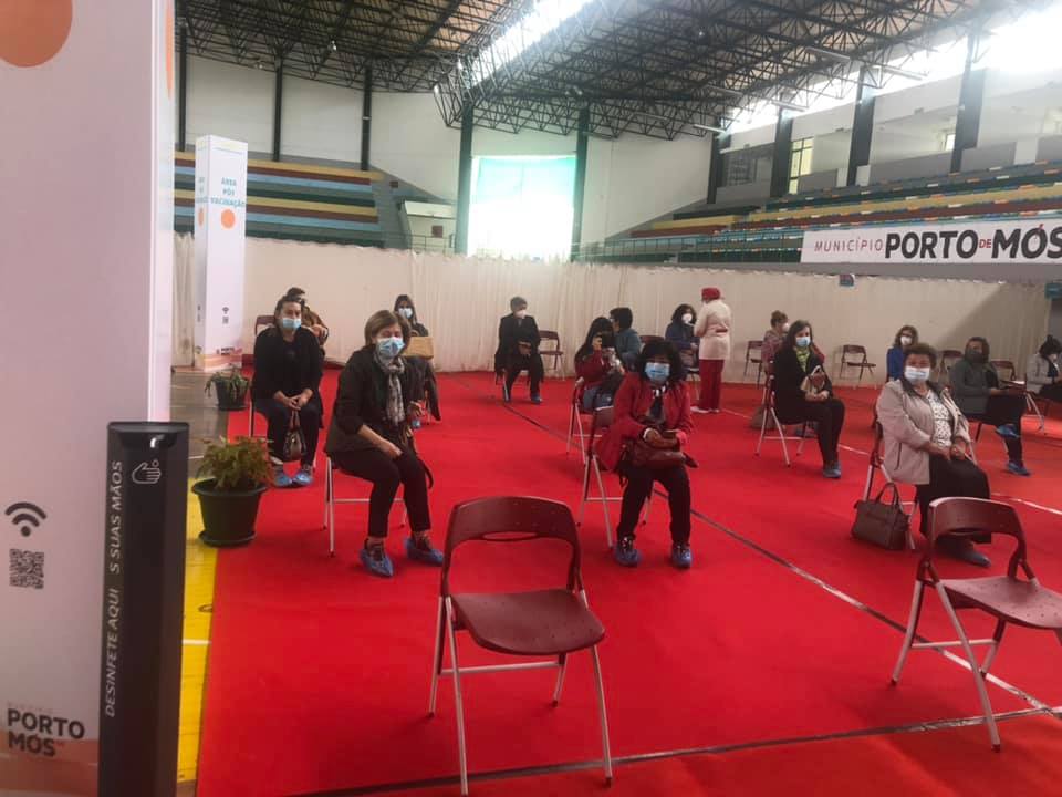 Pessoas de máscara aguardam sentadas em cadeiras afastadas num pavilhão desportivo. Na imagem, é visível a identificação do Município de Porto de Mós.