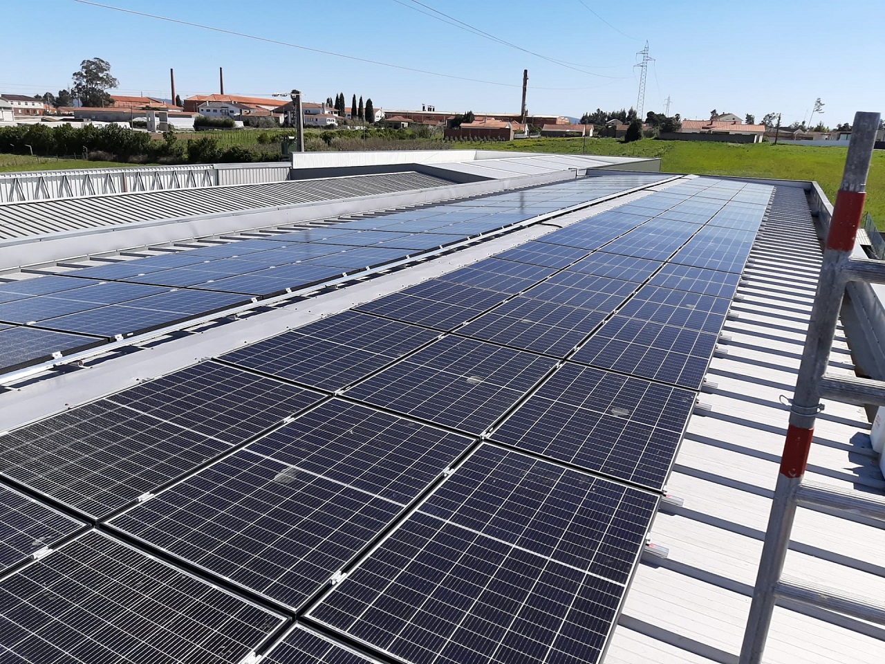 Painéis solares fotovoltaicos instalados na cobertura de um edifício. Ao fundo, veem-se casas, campos e uma linha elétrica.