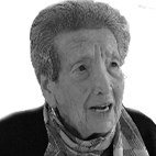 Retrato a preto e branco de uma pessoa idosa, com cabelo grisalho, casaco escuro e lenço ao pescoço. A pessoa olha para o lado e parece estar a falar.