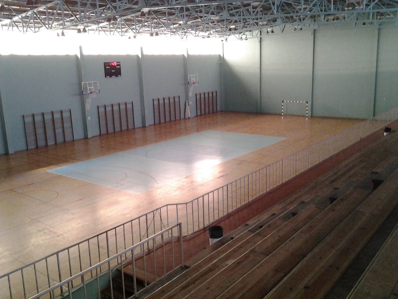 Vista de um pavilhão desportivo vazio, com campo marcado no piso, balizas, tabelas de basquetebol e bancadas de madeira. Não há pessoas visíveis.