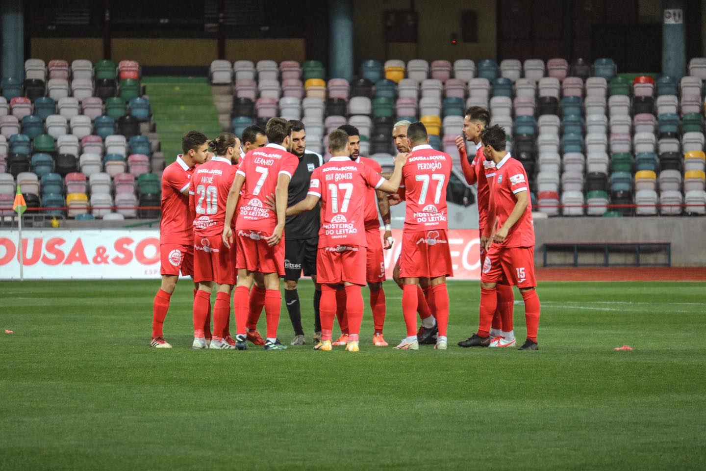 Um grupo de futebolistas de equipamento vermelho reúne-se no relvado em torno de um elemento vestido de preto. Nas camisolas são visíveis os nomes André, Leandro, Rui Gomes e Perdigão.