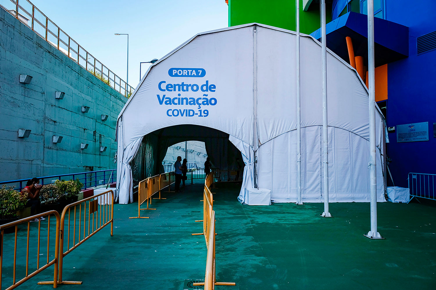 Entrada de um centro de vacinação contra a COVID-19, assinalada como «Porta 7». Grades metálicas orientam o acesso à tenda, onde se veem algumas pessoas.