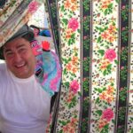 Um homem sorridente afasta uma cortina de tecido florido. Junto a ele, vê-se um boneco colorido.