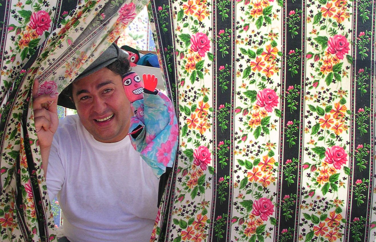 Um homem sorridente afasta uma cortina de tecido florido. Junto a ele, vê-se um boneco colorido.