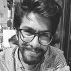 Retrato a preto e branco de um homem com óculos e barba, a olhar para a câmara. Veste uma camisola aos quadrados.