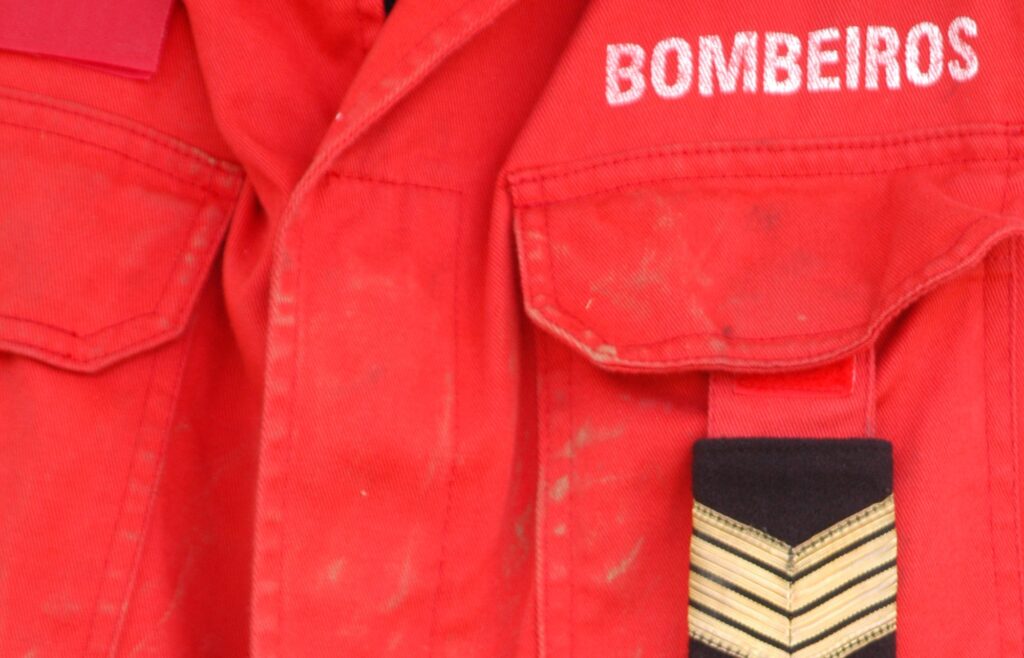 Grande plano de uma farda vermelha com a palavra «BOMBEIROS» e uma insígnia de graduação no bolso. Não são visíveis rostos nem elementos que permitam identificar uma pessoa.