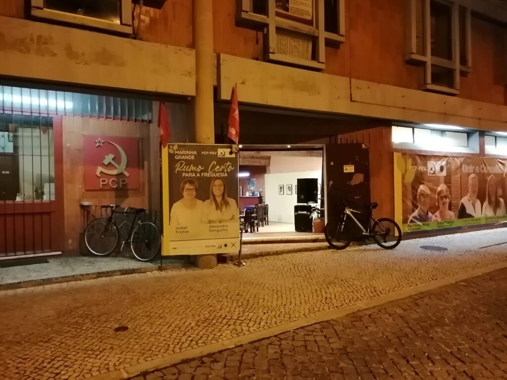 Fotografia noturna da fachada de um espaço do PCP na Marinha Grande. À entrada, um cartaz da CDU apresenta a candidatura «Rumo Certo para a Freguesia»; veem-se também bandeiras e bicicletas.