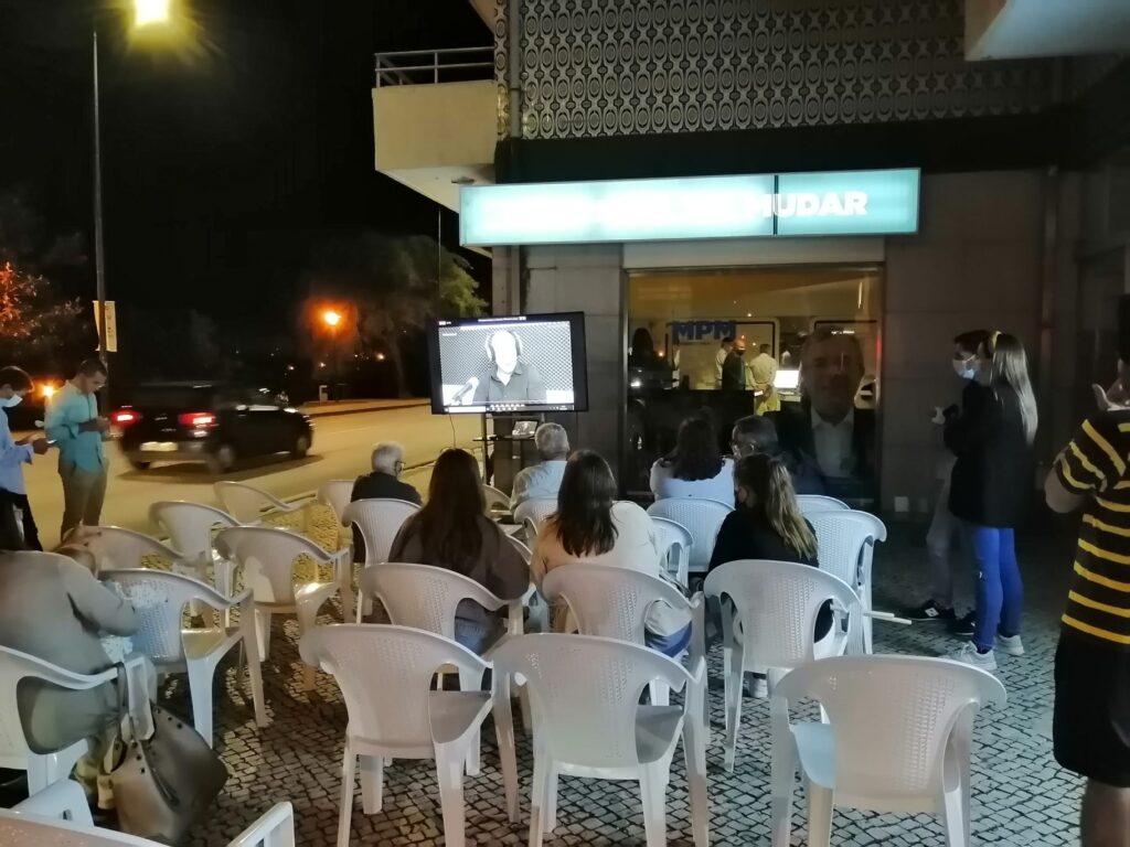 À noite, várias pessoas estão sentadas ao ar livre a assistir a uma pessoa num ecrã. A sessão decorre junto à entrada de um edifício.