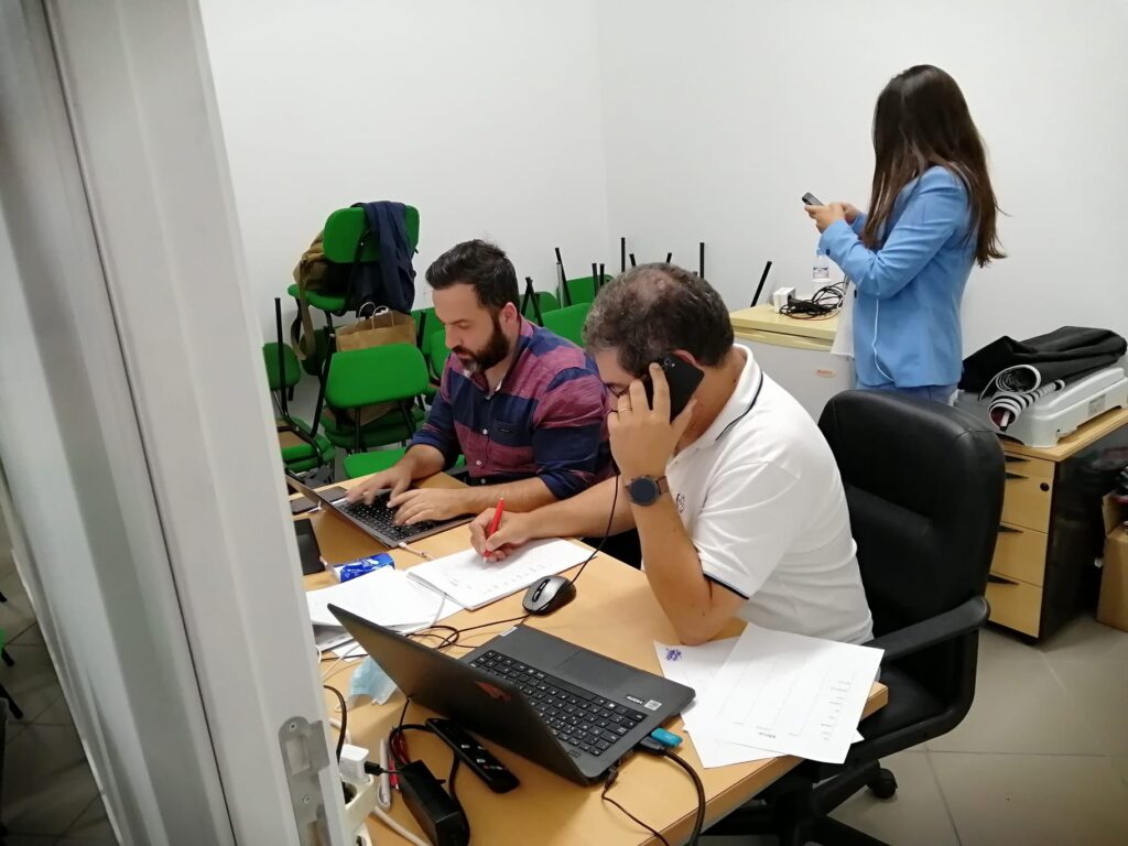 Três pessoas trabalham numa sala com computadores portáteis e documentos. Uma está ao telefone, outra usa o computador e a terceira consulta o telemóvel.