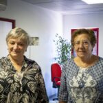 Duas mulheres posam lado a lado num espaço de escritório. Ao fundo, vêem-se cadeiras, uma planta e uma terceira pessoa parcialmente visível.