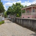 Uma entrada empedrada conduz a um portão metálico, junto a um edifício rosa com varanda. Muros e árvores rodeiam o espaço.