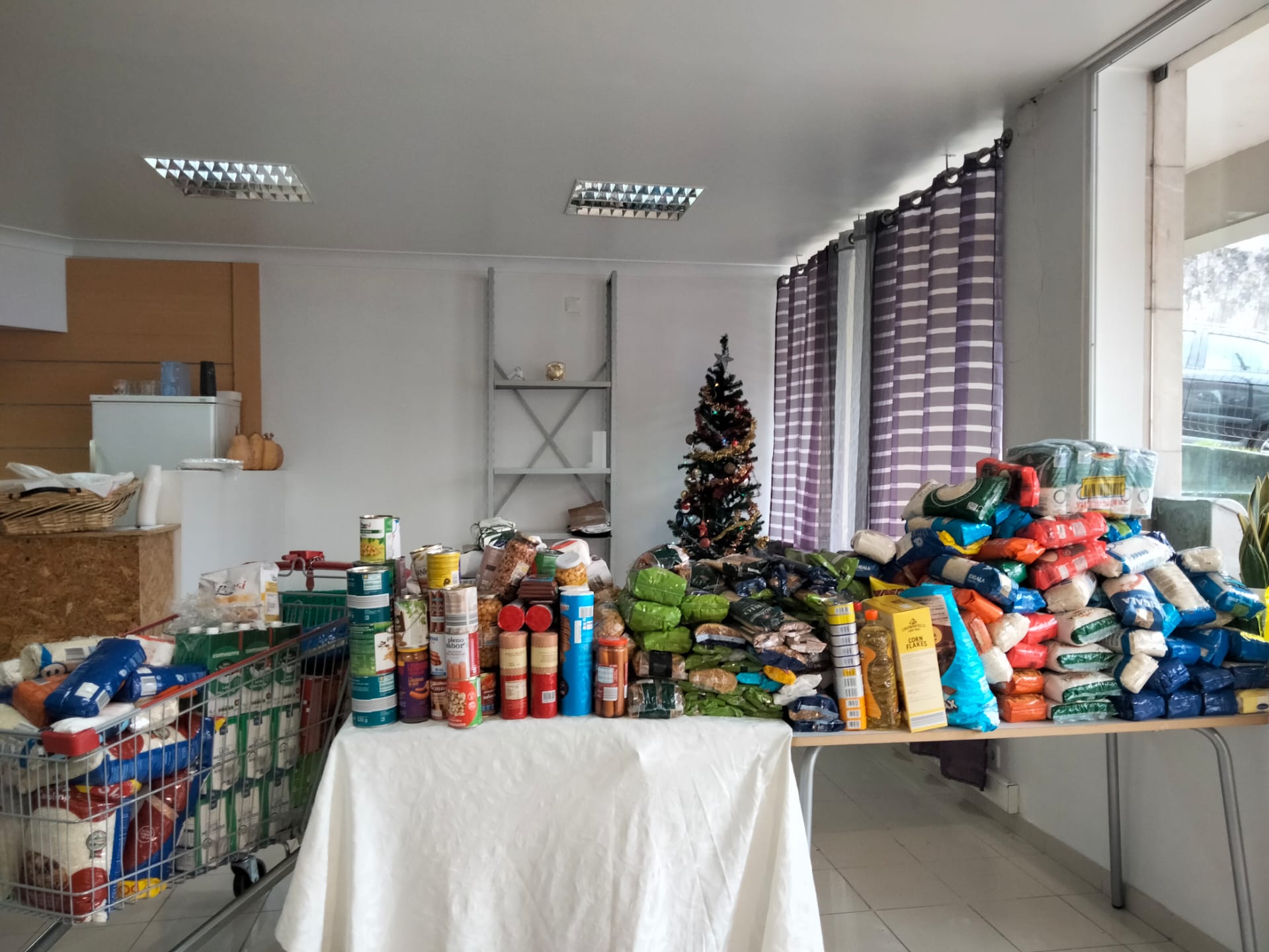 Uma sala contém grandes quantidades de alimentos embalados, incluindo arroz, conservas e cereais, dispostos sobre mesas e num carrinho de compras. Ao fundo, vê-se uma árvore de Natal decorada.
