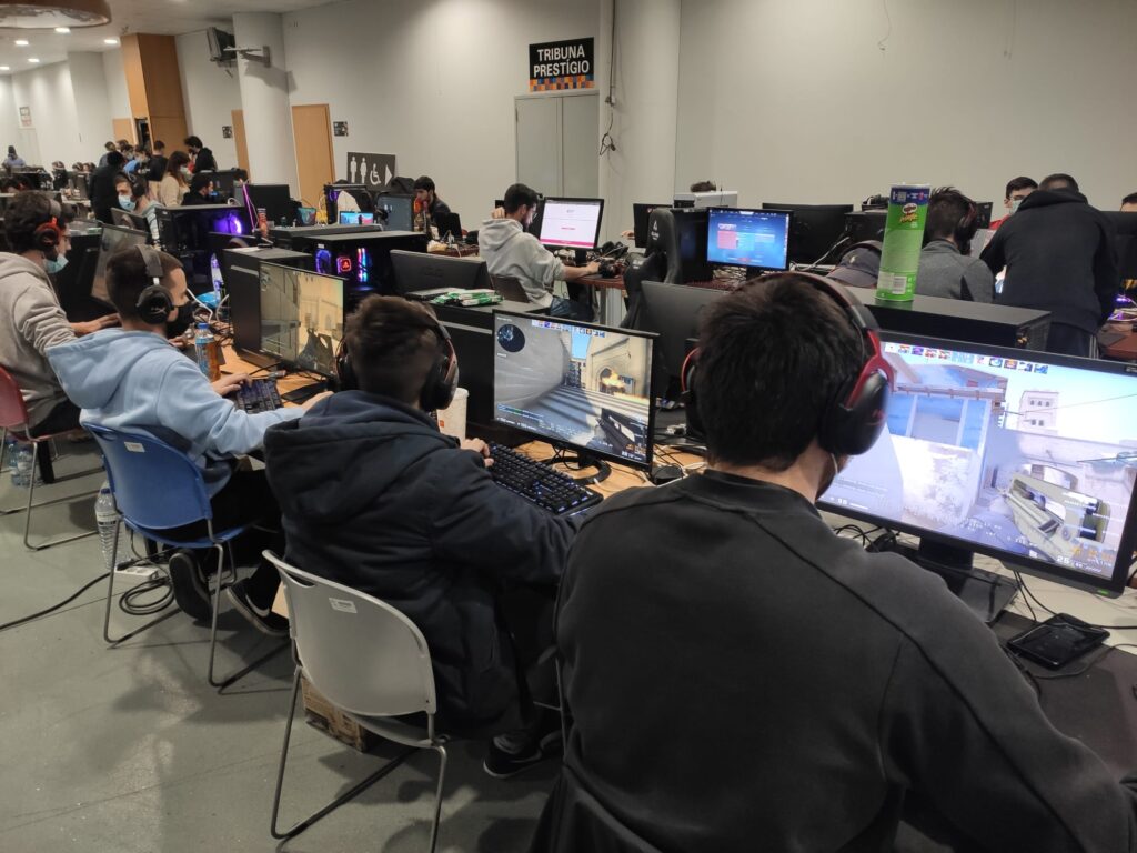 Várias pessoas jogam videojogos em computadores numa sala partilhada, usando auscultadores. Alguns ecrãs mostram um jogo de tiros; ao fundo, vê-se uma placa com a inscrição «Tribuna Prestigio».