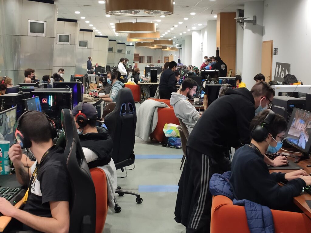 Uma sala ampla reúne várias pessoas a utilizar computadores, muitas com auscultadores e máscaras. Os postos estão dispostos em filas, com ecrãs e cadeiras de escritório.