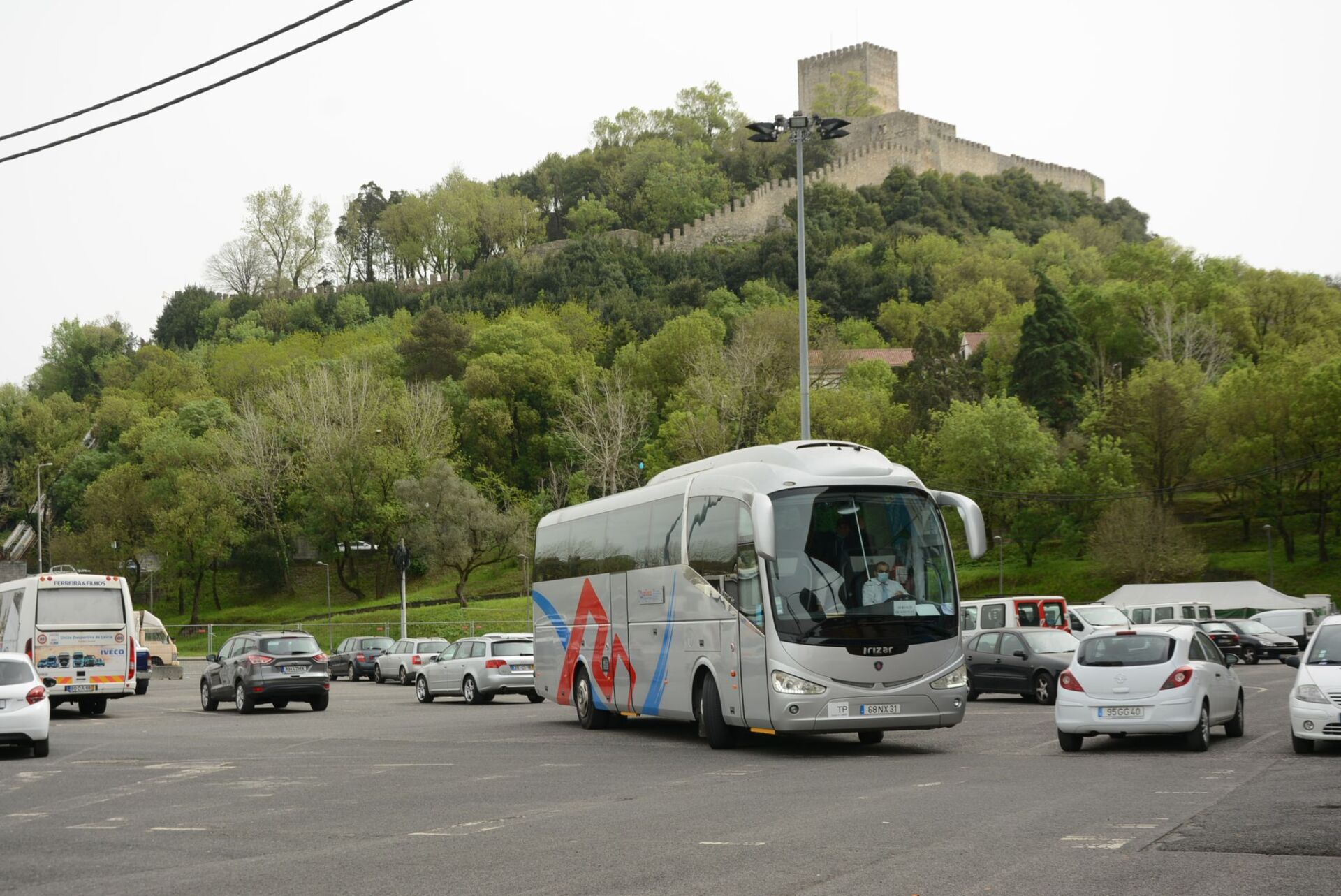 Um autocarro de turismo circula numa zona de estacionamento, com vários automóveis à volta. Ao fundo, vê-se uma fortificação com muralhas e uma torre no topo de uma colina arborizada.