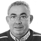Retrato a preto e branco de um homem de óculos, com cabelo curto grisalho. Veste uma camisola escura sobre uma camisola de padrão aos quadrados.