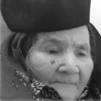 Fotografia a preto e branco de uma mulher idosa em grande plano. Usa um chapéu escuro e um lenço ao pescoço.