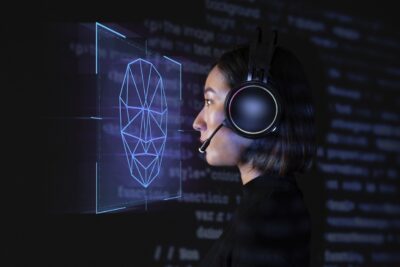 Uma pessoa com auscultadores e microfone observa uma representação digital de um rosto formada por linhas geométricas. A imagem sugere o tema do reconhecimento facial, mas não mostra uma identificação concluída.