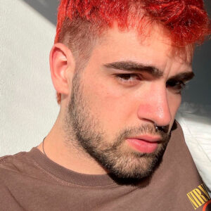 Retrato aproximado de uma pessoa com cabelo vermelho, barba e piercing no nariz. Usa uma camisola castanha e está iluminada pelo sol.