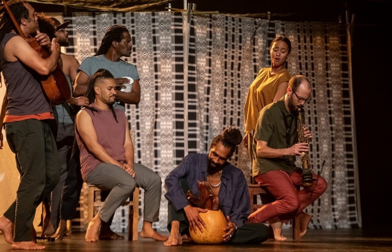 Um grupo de artistas atua em palco, com canto, dança e instrumentos musicais. Vêem-se um instrumento de sopro e instrumentos de percussão.