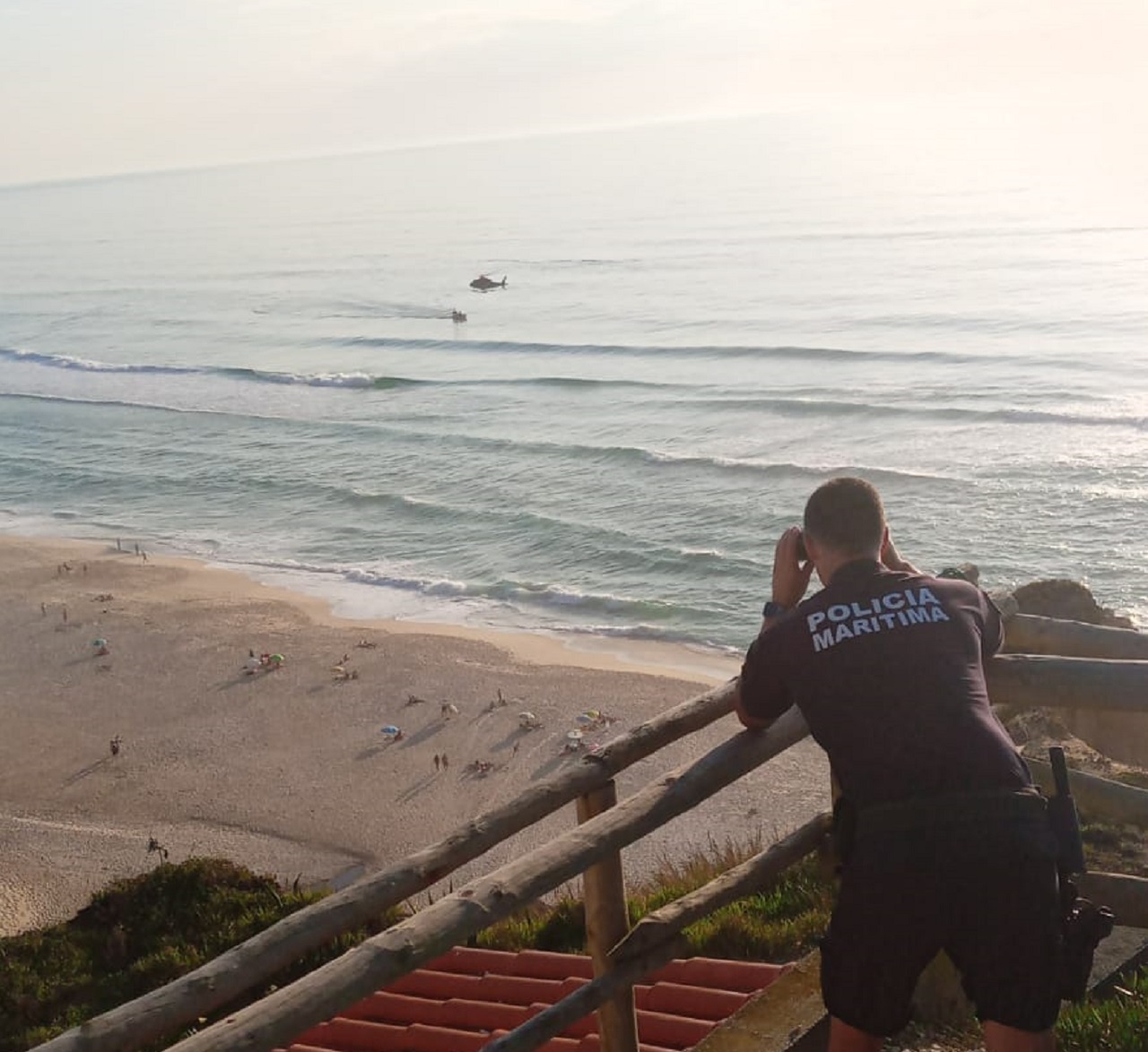 Agente da Polícia Marítima com binóculos acompanha as b uscas no mar em que participa um helicóptero