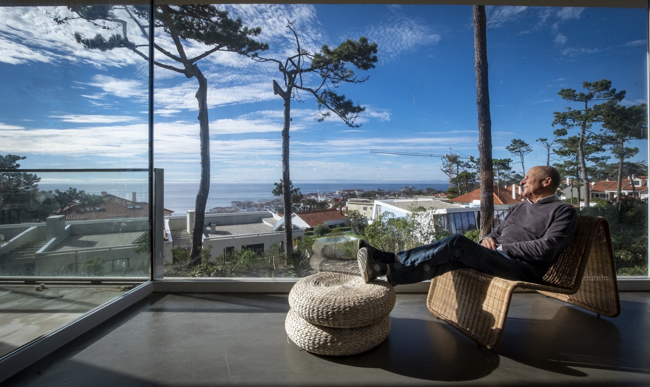 Foto de Gonçalo Byrne sentado numa poltrona no interior da sua casa a olhar o mar de são pedro de moel