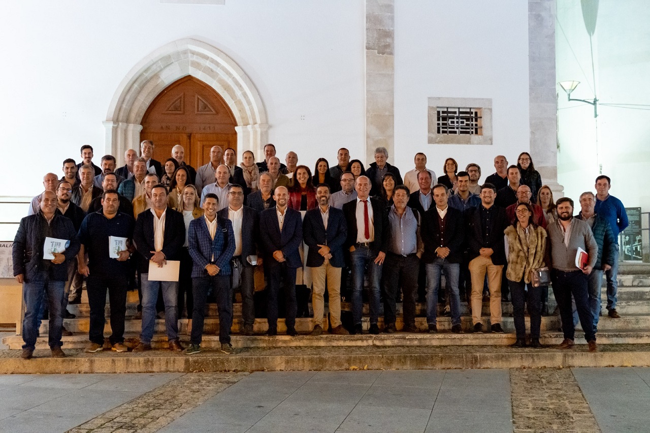 Um grupo numeroso posa para uma fotografia nas escadas à entrada de um edifício de fachada branca e porta em arco. Algumas pessoas seguram documentos.