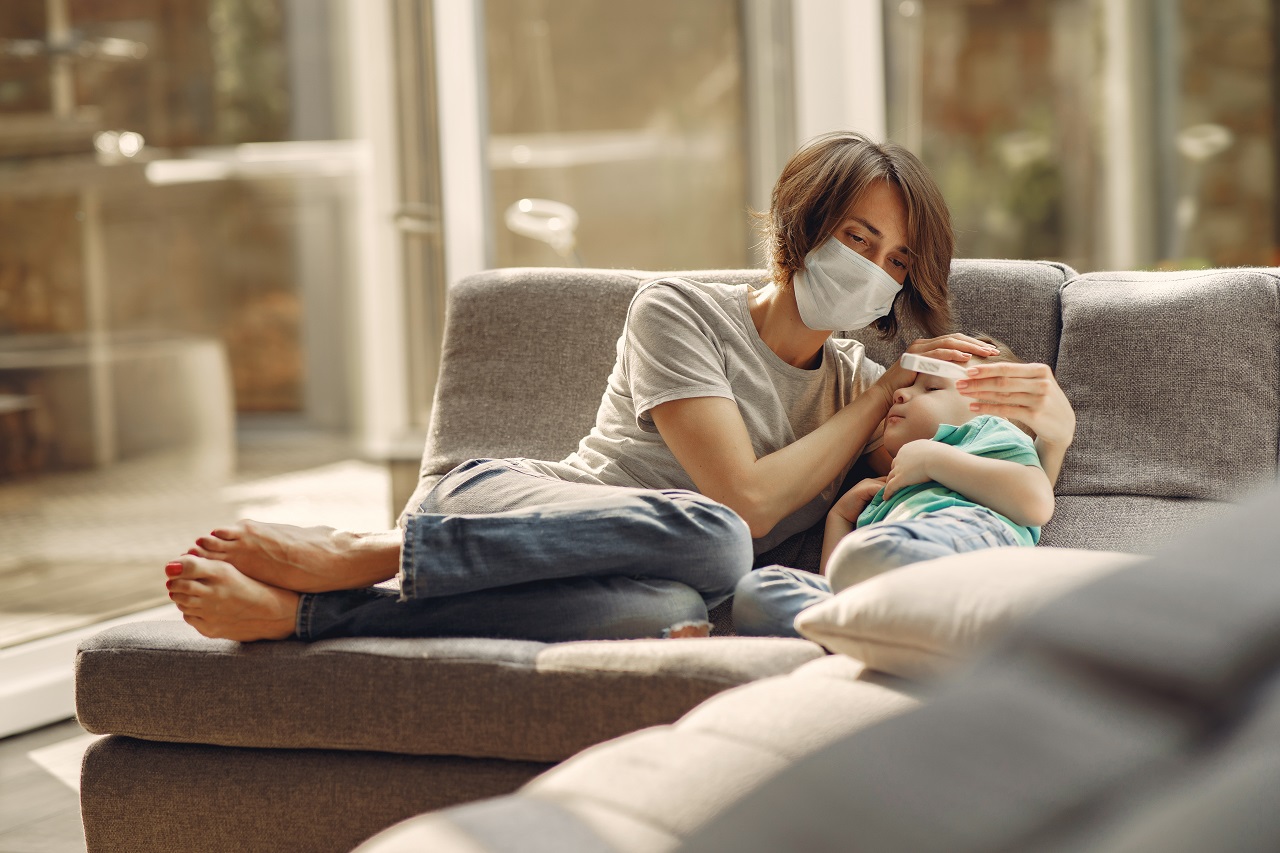 Mulher com máscara e um termómetro na mão está sentada no sofá com um menino, com ar adoentado