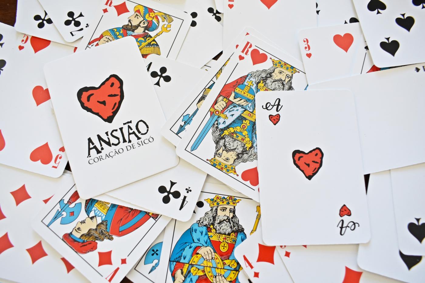 Várias cartas de jogar estão espalhadas e sobrepostas. Uma das cartas apresenta um coração vermelho e o texto «ANSIÃO CORAÇÃO DE SICÓ».