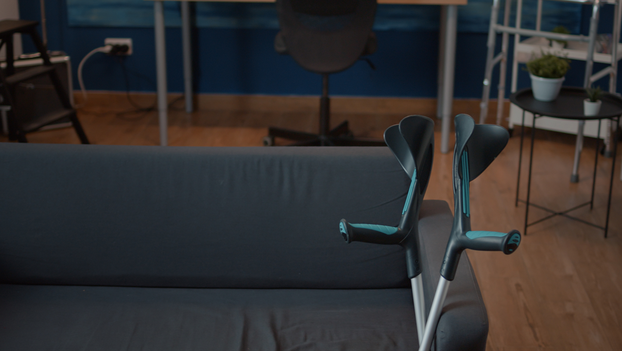 Fotografia de um par de muletas encostadas a um sofá cinzento em primeiro plano, numa sala, onde se vê algum mobiliário