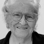 Retrato a preto e branco de uma mulher idosa, sorridente e com óculos. O fundo é claro e não há texto visível.